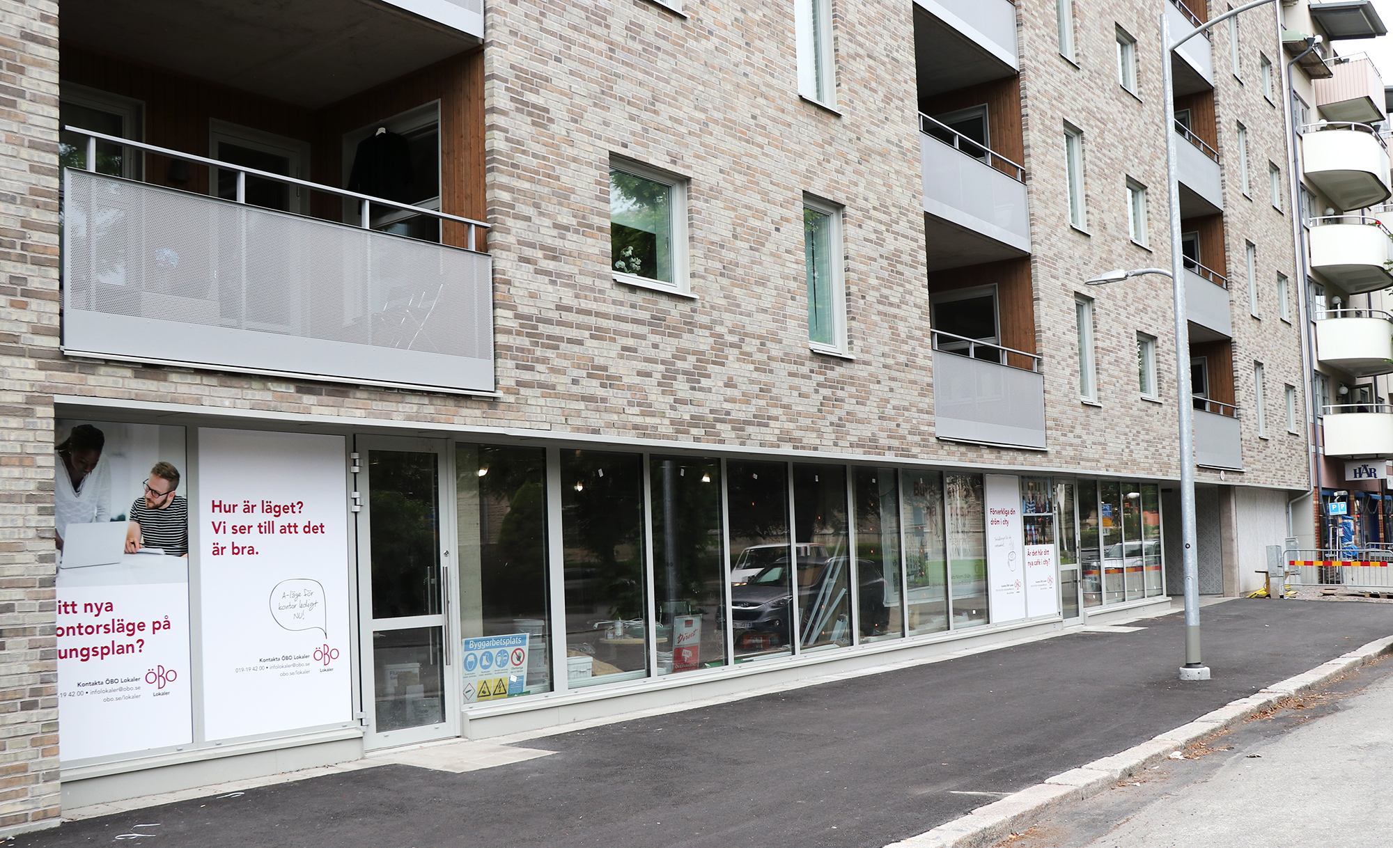 Lång husfasad med stora glasfönster i bottenplan och balkonger på de övre våningarna. Byggnaden är klädd i ljus tegel och gatan framför är nyasfalterad.