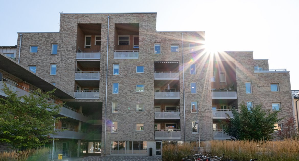 Flerbostadshus i tegel med flera våningar och balkonger. Solen lyser in från höger och träd samt cyklar syns i förgrunden.